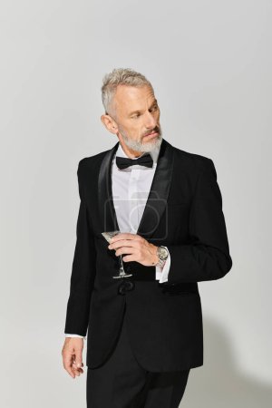 gut aussehender reifer Mann im eleganten Smoking mit Bart hält Martini mit Oliven und schaut weg