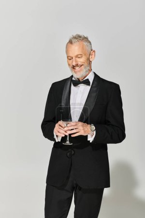 fröhlich gut aussehender reifer Mann im schicken Smoking mit grauem Bart lächelnd und Martini mit Oliven in der Hand
