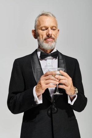 attraktiver bärtiger älterer Mann im eleganten Smoking mit Fliege, der ein Glas Martini mit Oliven hält