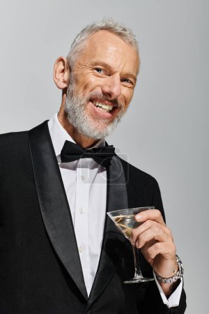 fröhlich gut aussehender reifer Mann im eleganten Smoking hält Glas Martini in der Hand und lächelt in die Kamera