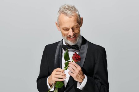 attractive joyful mature man with beard in elegant tuxedo holding red rose and smiling happily