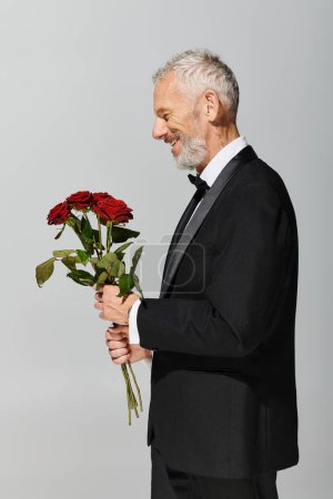 cheerful good looking mature man in voguish black tuxedo holding red roses bouquet in hands