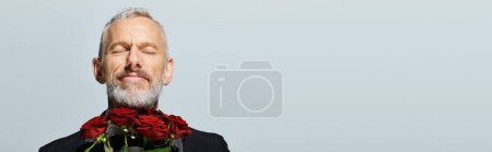 joyful good looking mature man in chic tuxedo holding red roses bouquet and smiling with closed eyes