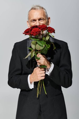 fröhlich gut aussehender, reifer Mann im modischen Smoking, rote Rosen in der Hand und glücklich lächelnd