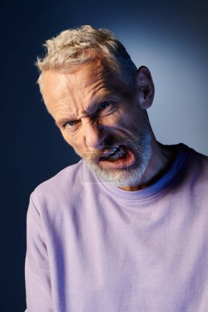 schöner bärtiger älterer Mann mit Accessoires in stylischer Kleidung, der vor blauem Hintergrund aktiv grimmig wirkt