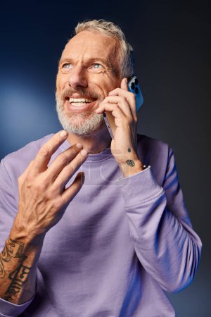 cheerful bearded trendy mature man in purple stylish sweatshirt talking by phone on blue backdrop