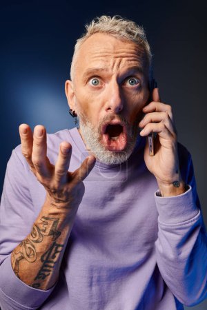 handsome fashionable mature man in purple stylish sweatshirt grimacing while talking by phone