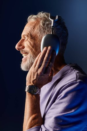 joyful attractive mature man with gray beard and headphones in purple sweatshirt enjoying music