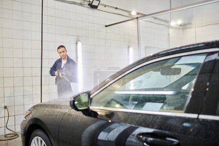 attractive dedicated professional in blue uniform using hose to wash modern black car in garage