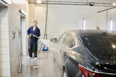 good looking devoted serviceman in blue uniform with collected hair using hose to wash black car