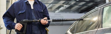 vista recortada del profesional trabajador duro en uniforme azul usando la manguera para lavar el coche negro, bandera