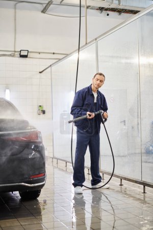 atractivo trabajador profesional en uniforme azul con el pelo recogido usando manguera para lavar el coche