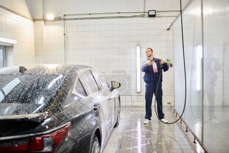 schöner professioneller Arbeiter in blauer Uniform, der Seife benutzt, um sein schwarzes modernes Auto in der Garage zu waschen