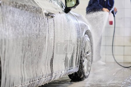 przycięty widok dedykowanego ciężko pracującego człowieka w niebieskim mundurze myjącego samochód z mydłem w garażu