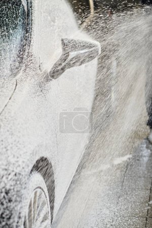 abgeschnittene Ansicht des engagierten professionellen Arbeiters, der Schlauch hält und schwarzes modernes Auto mit Seife wäscht
