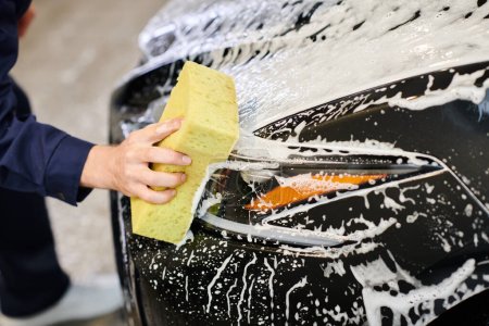 cropped view of dedicated hard working man in uniform washing black car with sponge and soap
