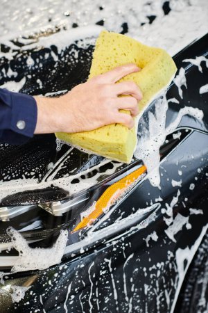 cropped view of dedicated specialist in blue uniform using soapy sponge to wash black modern car