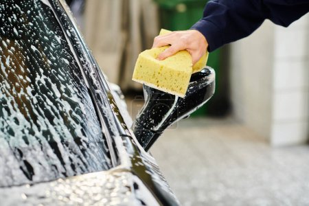 cropped view of hard working professional serviceman in uniform using sponge to wash car in garage