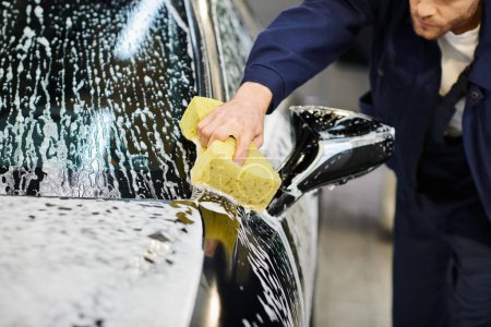 przycięty widok dedykowanego profesjonalnego pracownika w niebieskim mundurze za pomocą gąbki mydlanej do mycia czarnego samochodu