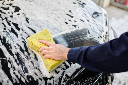 przycięty widok oddanego profesjonalnego pracownika w niebieskim mundurze mycie czarny samochód z gąbką mydlaną
