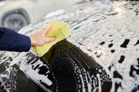 przycięty widok dedykowanego profesjonalnego pracownika w niebieskim mundurze za pomocą gąbki do mycia czarnego samochodu