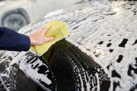 vista recortada de trabajador profesional dedicado en uniforme azul usando esponja para lavar coche negro