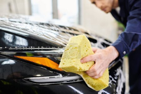 focus on soapy yellow sponge in hands of blurred attractive serviceman washing black modern car