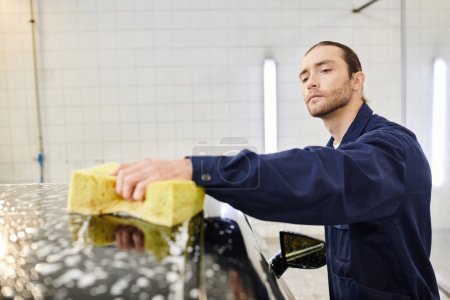 attractive devoted serviceman in comfy blue uniform washing car with soapy sponge while in garage