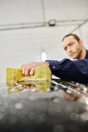 focus on yellow soapy sponge in hands of blurred handsome man in uniform washing black car