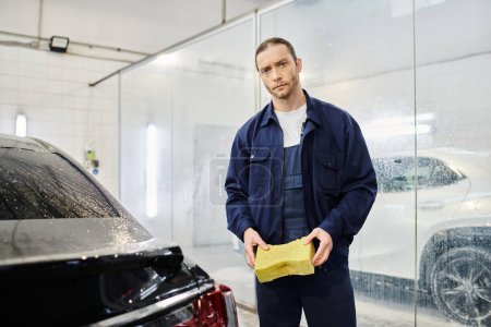 handsome professional worker in blue uniform holding soapy sponge and looking at camera in garage