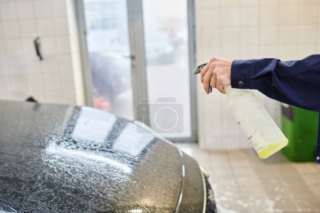 vue recadrée du travailleur professionnel enthousiaste en uniforme en utilisant pulvérisateur avec de l'eau pour nettoyer la voiture