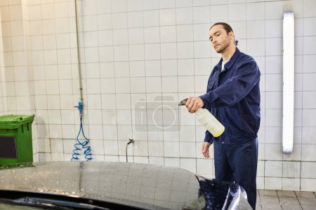 gut aussehender, hart arbeitender Mann mit gesammelten Haaren in blauer Uniform mit Pulverisierer, um Auto zu reinigen