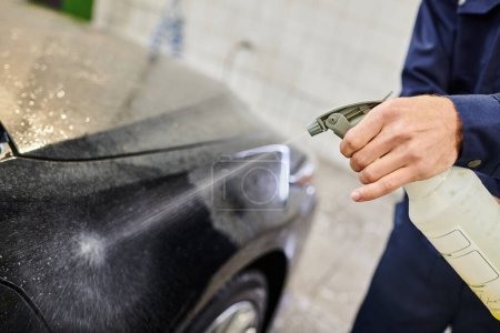 cropped view of hard working professional in uniform using pulverizer to clean black modern car