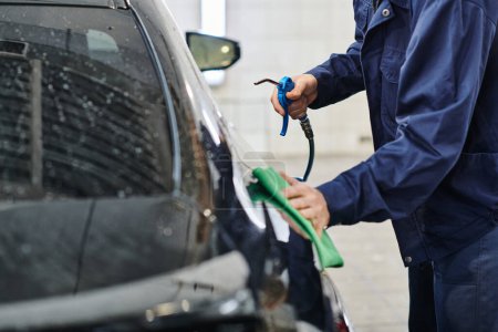 cropped view of dedicated professional worker in uniform holding hose and cleaning car with rag