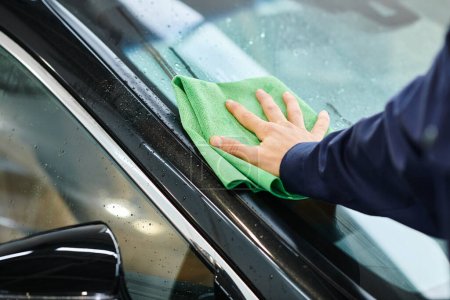 cropped view of hard working specialist in uniform cleaning black car with green rag in garage