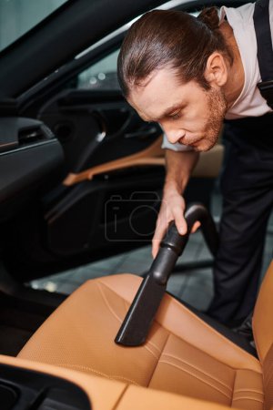 good looking hard working serviceman in uniform using manual vacuum cleaner in car in garage