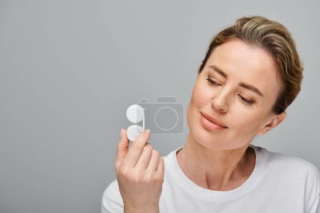 good looking jolly woman with blonde hair holding contact lenses and looking away on gray backdrop