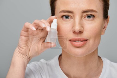 cheerful good looking woman holding eye drops and looking straight at camera on gray backdrop