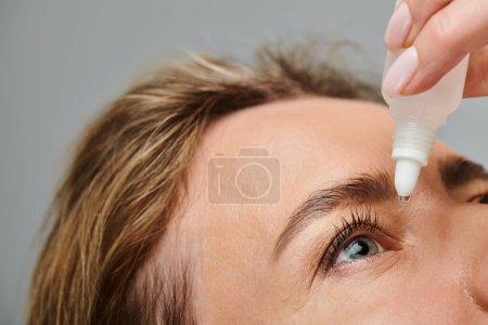 close up of attractive female model with blonde hair putting in eye drops while on gray backdrop