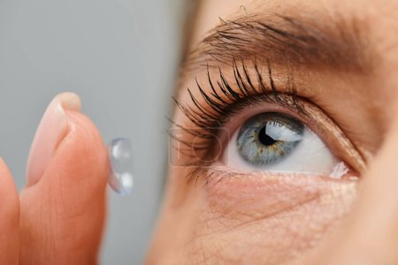 Nahaufnahme einer gut aussehenden Frau, die aufmerksam ihre Kontaktlinse trägt, während sie auf grauem Hintergrund posiert