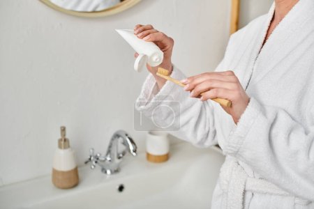 abgeschnittene Ansicht einer erwachsenen Frau im kuscheligen Bademantel, die Zahnpasta auf ihre Bürste in der Nähe des Spiegels legt