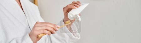 cropped view of adult woman in comfy bathrobe putting tooth paste on her brush near mirror, banner