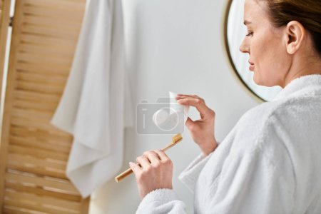 gut aussehende blonde Frau im Bademantel beim Zähneputzen vor dem Spiegel in ihrem Badezimmer