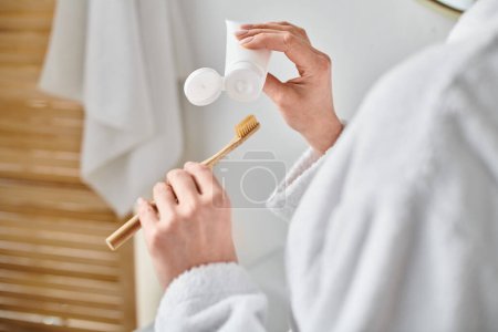 abgeschnittene Ansicht einer erwachsenen Frau im kuscheligen Bademantel, die Zahnpasta auf ihre Bürste in der Nähe des Spiegels legt