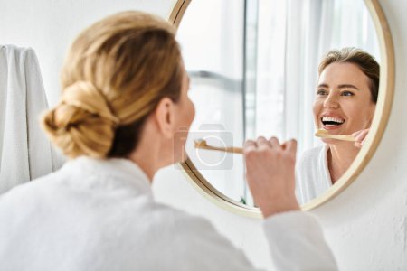 attractive joyous woman with blonde hair in bathrobe brushing her teeth and looking at mirror