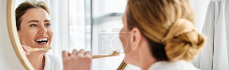 atractiva mujer alegre con cabello rubio en albornoz cepillándose los dientes y mirando al espejo