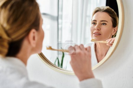 good looking blonde woman in bathrobe brushing her teeth in front of mirror in her bathroom