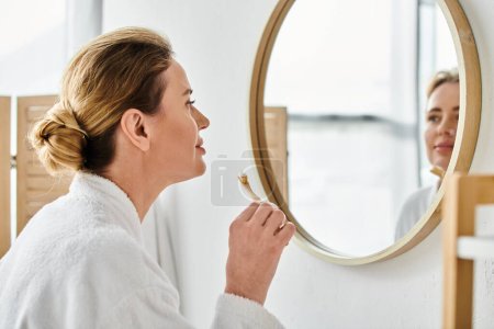 Photo for Good looking blonde woman in bathrobe brushing her teeth in front of mirror in her bathroom - Royalty Free Image