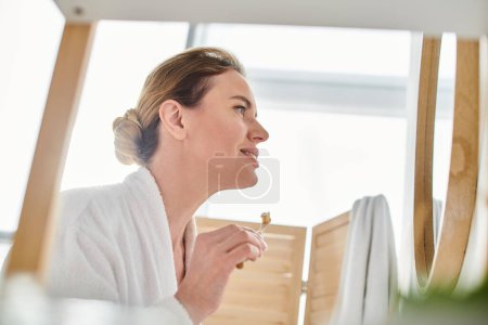 Photo for Appealing joyful woman with blonde hair in bathrobe brushing her teeth and looking at mirror - Royalty Free Image