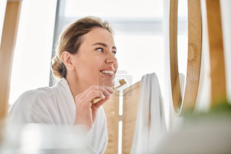 appealing joyful woman with blonde hair in bathrobe brushing her teeth and looking at mirror